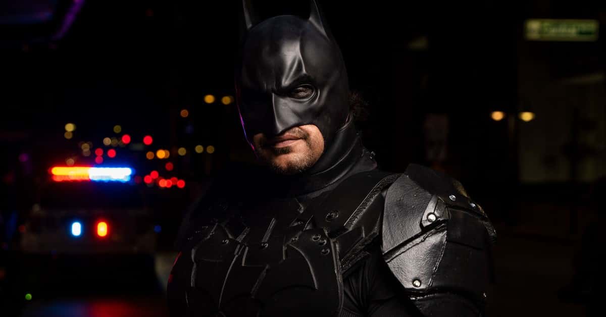 Batman cosplayer in detailed costume at a night event with police lights in the background.