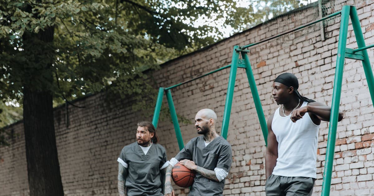 Three men in a prison yard, one holding a basketball, alongside a brick wall.