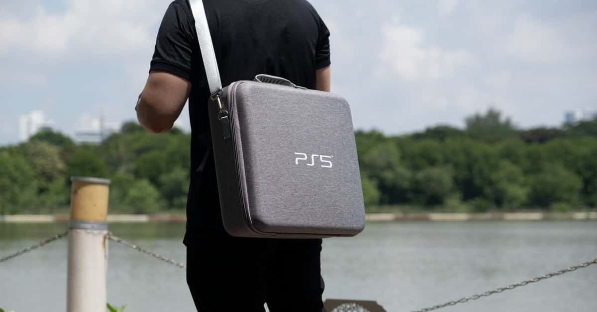 A man carries a PS5 bag by a lake, surrounded by greenery in a park setting.