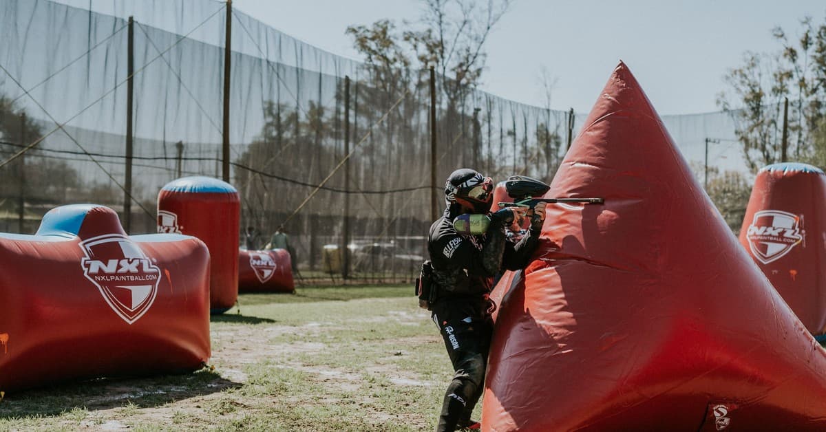 Paintball player strategizing behind inflatable cover during a competition on an outdoor field.