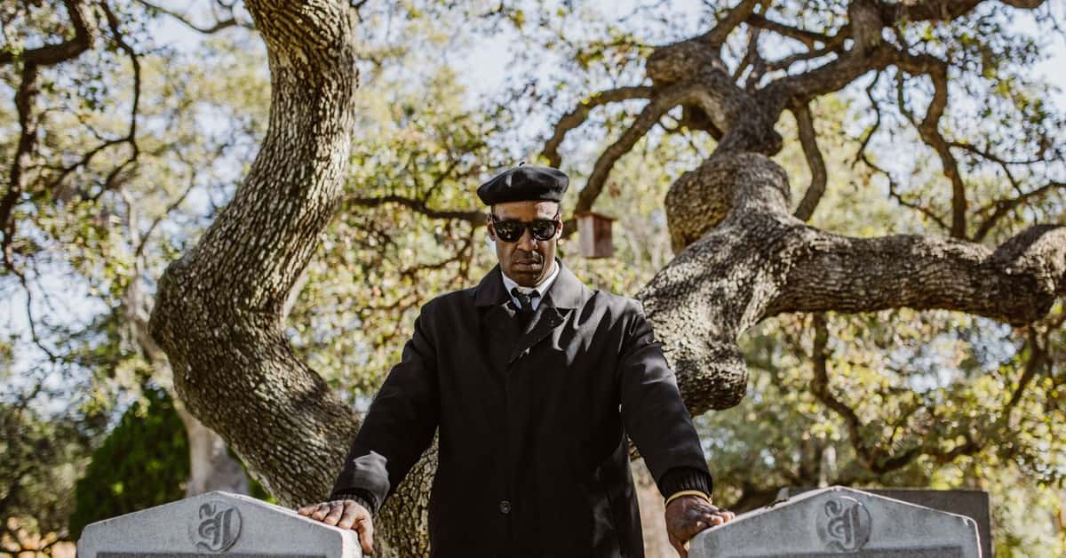 A man wearing a black coat and sunglasses stands by tombstones in a cemetery, surrounded by trees.
