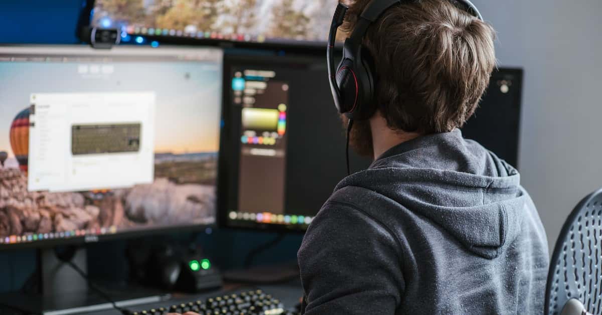 Back view anonymous male in casual clothes and headphones using modern computer with big monitors and typing on backlit keyboard
