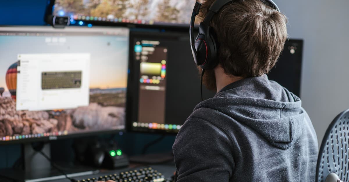 Back view anonymous male in casual clothes and headphones using modern computer with big monitors and typing on backlit keyboard