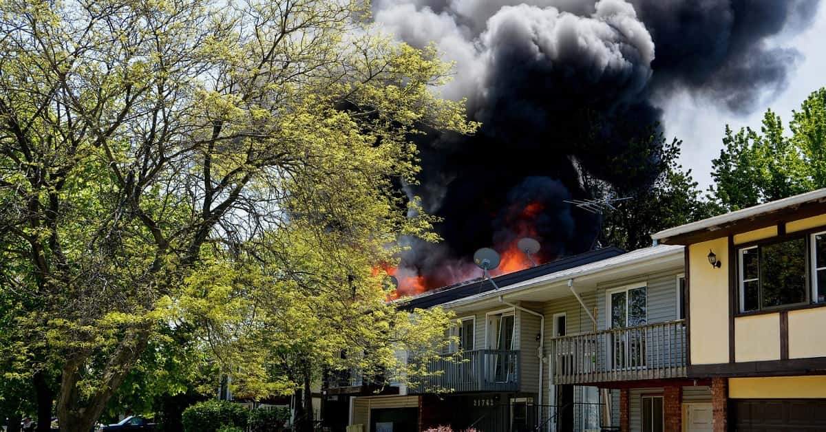 Dramatic house fire with thick smoke rising in a suburban neighborhood, surrounded by trees.