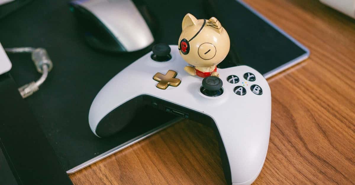 A charming cat figurine sits atop a gaming controller, beside a computer mouse, on a wooden desk.