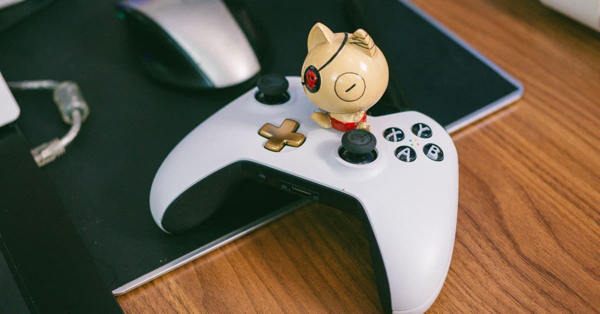 A charming cat figurine sits atop a gaming controller, beside a computer mouse, on a wooden desk.