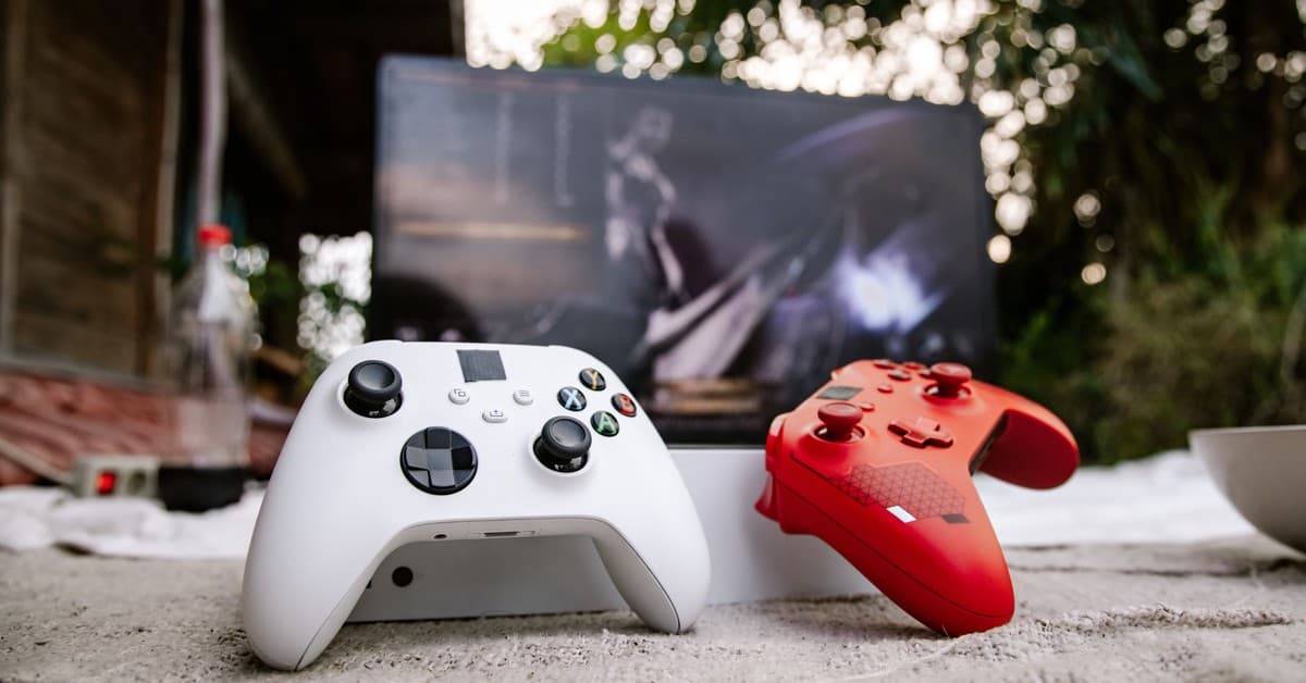 White and red Xbox wireless controllers set outdoors. A computer screen is blurred in the background.