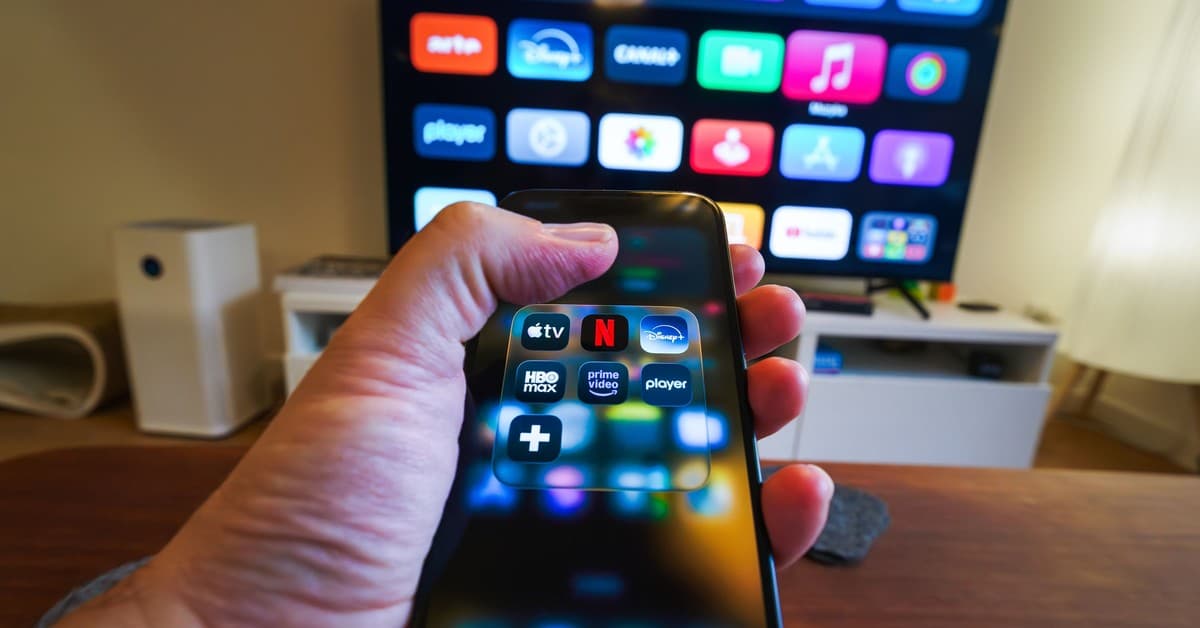 Close-up of a hand holding a phone displaying streaming apps in front of a TV with multiple app icons.