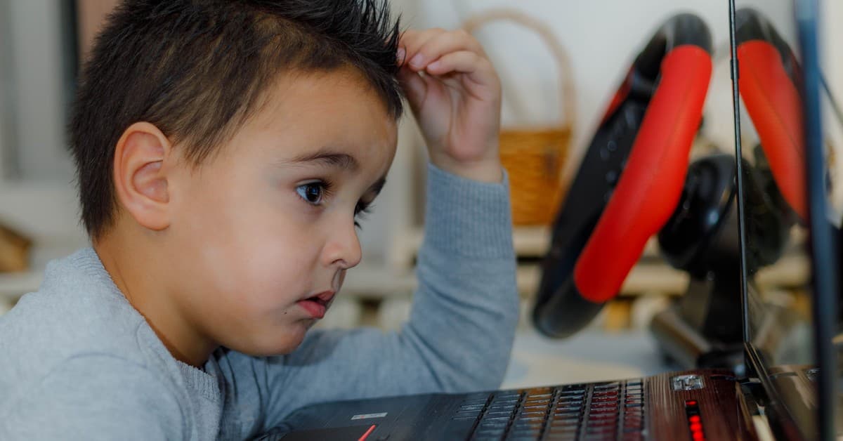 Child intensely focused on computer gaming with racing wheel accessory at home.