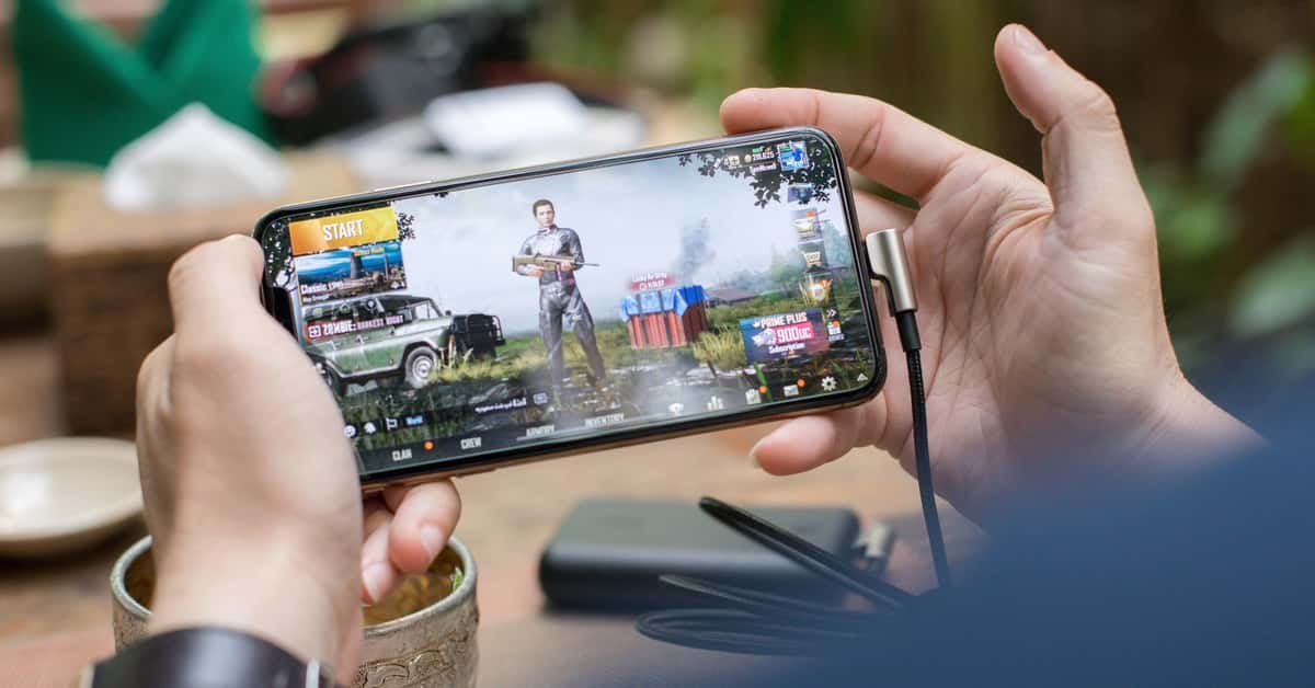 Person playing PUBG on smartphone outdoors with charging cable attached.