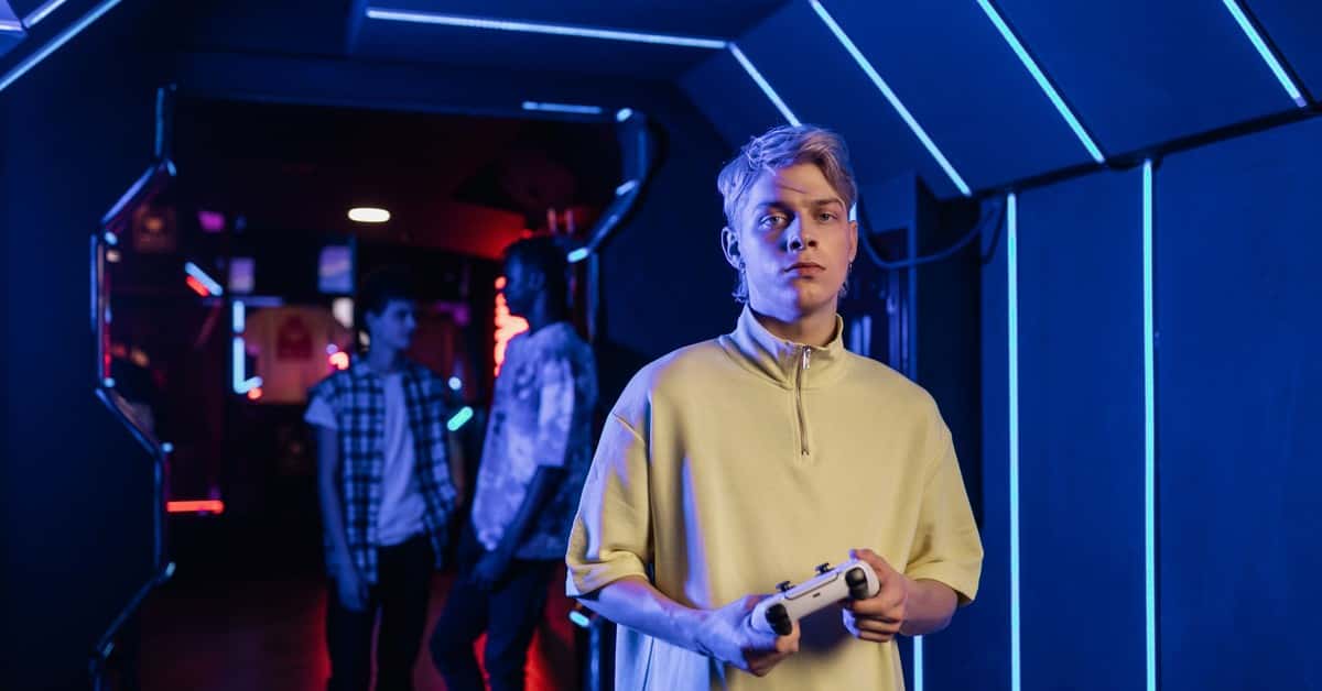 Serious young man holding a game controller in a neon-lit arcade room with friends in the background.