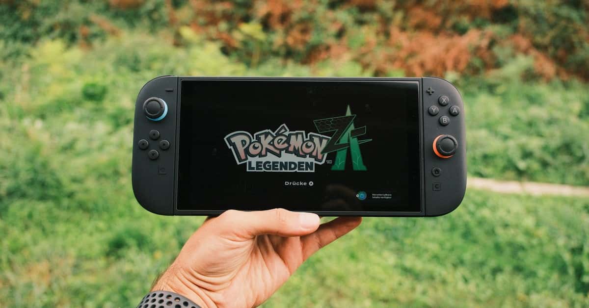 A hand holding a handheld gaming console displaying the Pokémon Legends game screen outdoors.