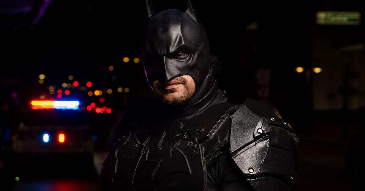 Batman cosplayer in detailed costume at a night event with police lights in the background.