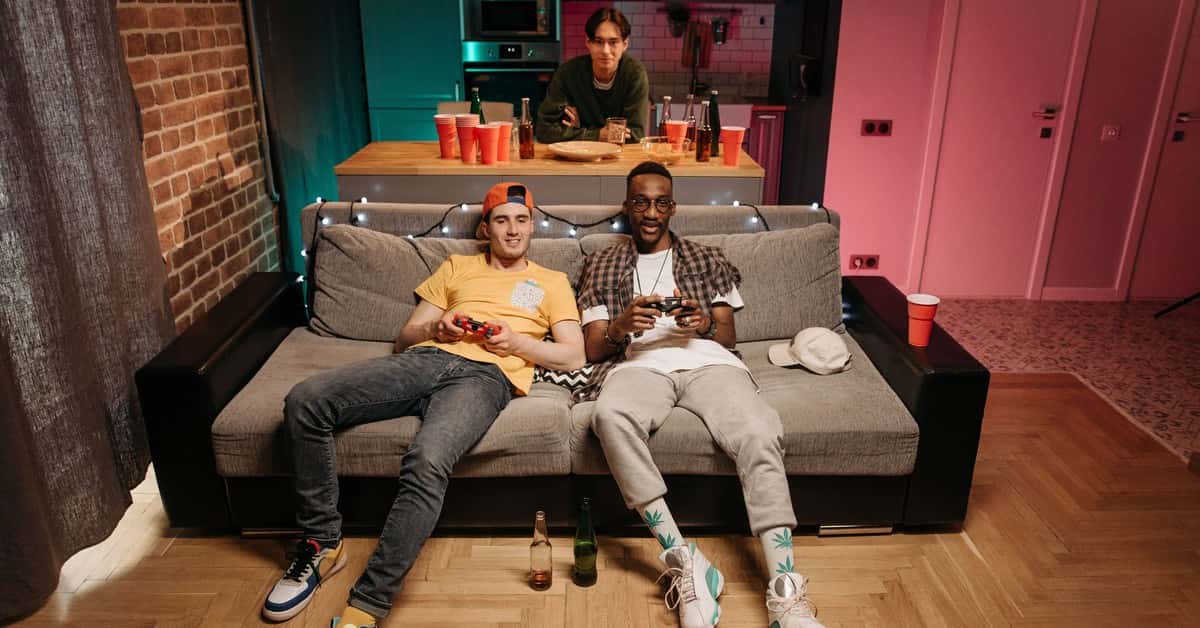 Three friends enjoying a gaming night in a cozy living room with colorful lights and drinks.