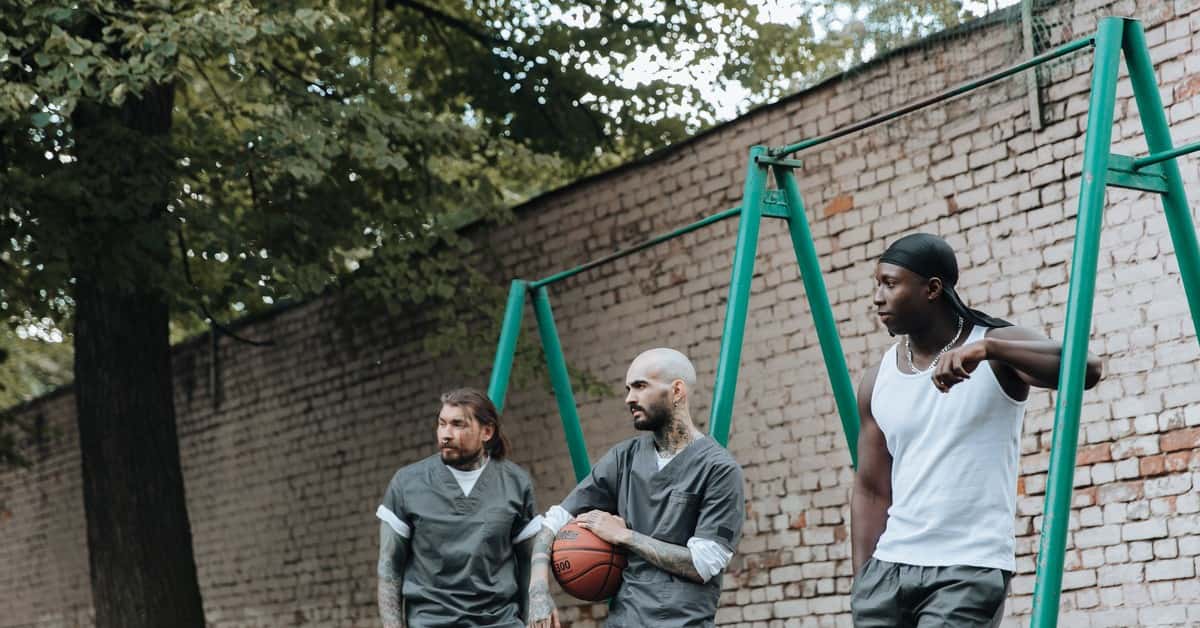 Three men in a prison yard, one holding a basketball, alongside a brick wall.
