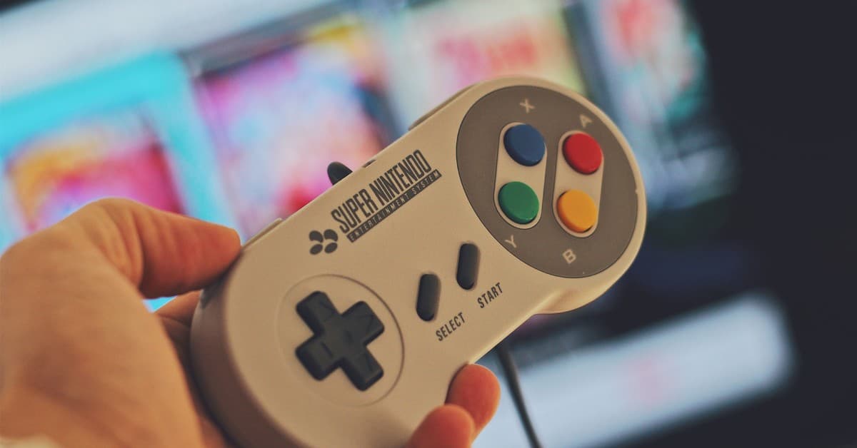 Hand holding a classic Super Nintendo game controller indoors with a blurred screen in the background.