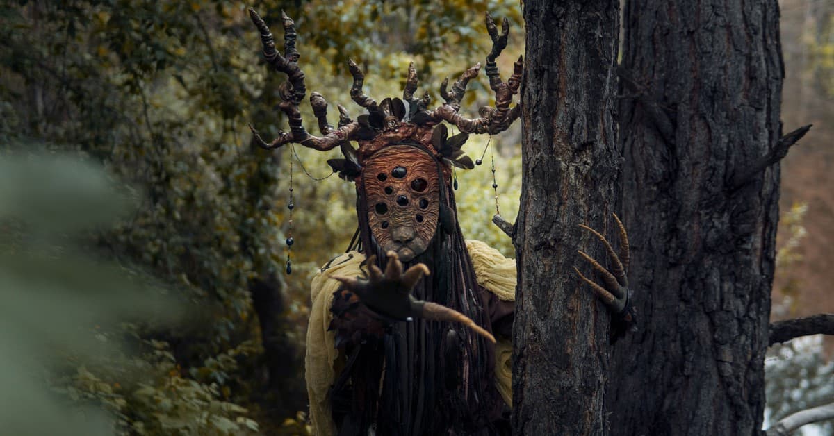 A mythical creature in the forest wearing a mask and antlers, hidden behind trees.