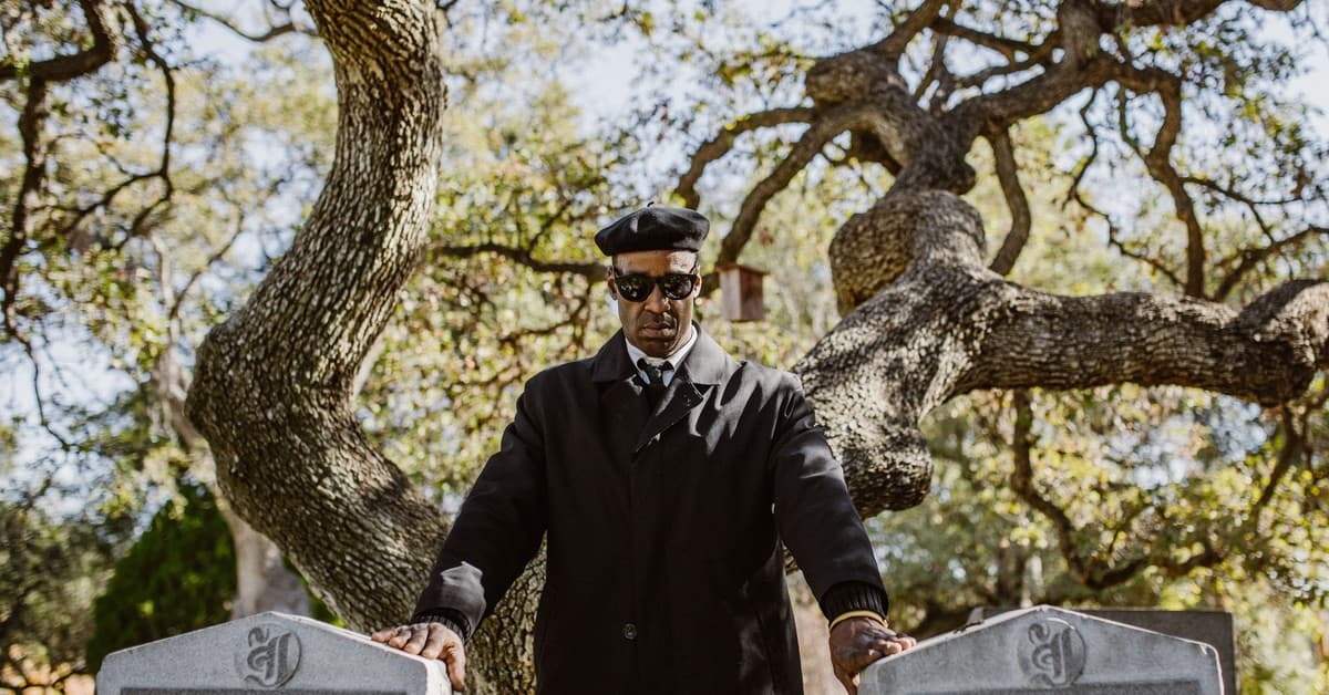 A man wearing a black coat and sunglasses stands by tombstones in a cemetery, surrounded by trees.
